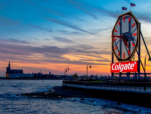 deniz kıyısı, arka planda kırmızı yelkovanlı büyük bir saatin altında Colgate logosu